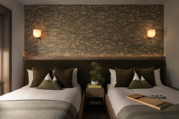 A warmly lit, compact twin bedroom with two beds arranged symmetrically against a dark upholstered full-width headboard. The room features layered white and olive bedding, textured grey throws, dark timber-toned bedside furniture, matching wall sconces, and a dramatic mottled green-grey feature wall behind the beds. A decorative game board rests on the right bed, and a small plant sits on the central nightstand.
