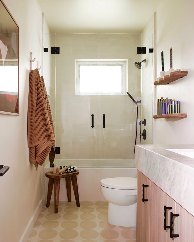 Bright, compact bathroom with a glass-enclosed shower over a bathtub, wall-mounted toilet, and a timber vanity with a pale stone benchtop. The room features cream wall tiles, patterned circular floor tiles, black hardware, wooden shelves, a rust-coloured towel, and playful colourful accessories.