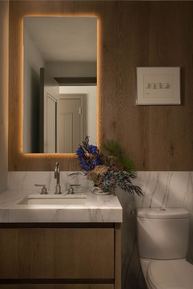 A compact, warmly lit bathroom featuring a floating dark timber vanity with an integrated white marble-look benchtop and backsplash, undermount sink, polished metal tapware, and a white toilet. A large rectangular backlit mirror is mounted on a dark timber wall panel, with framed artwork and a small floral arrangement adding decorative detail.