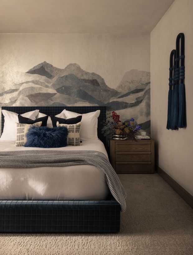 A refined bedroom with a deep navy upholstered bed and matching tiled headboard, layered white and grey bedding, and a vivid navy accent cushion. The room features a large monochrome mountain mural behind the bed, a dark timber bedside chest, decorative floral arrangement, beige carpet, and a sculptural navy wall hanging. The overall scheme is moody, luxurious, and contemporary with strong blue-grey contrast.