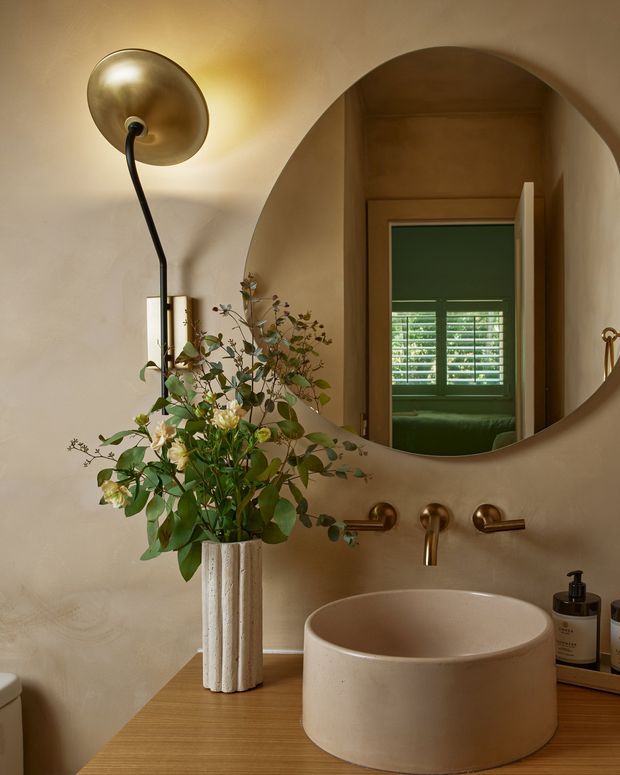 Warm, refined bathroom vanity featuring a round vessel basin on a timber counter, brushed brass wall-mounted tapware, a large rounded-arch mirror, and a sculptural brass wall light. A fluted white ceramic vase with greenery adds a natural decorative accent. The mirror reflects a green-toned adjoining room with plantation shutters.
