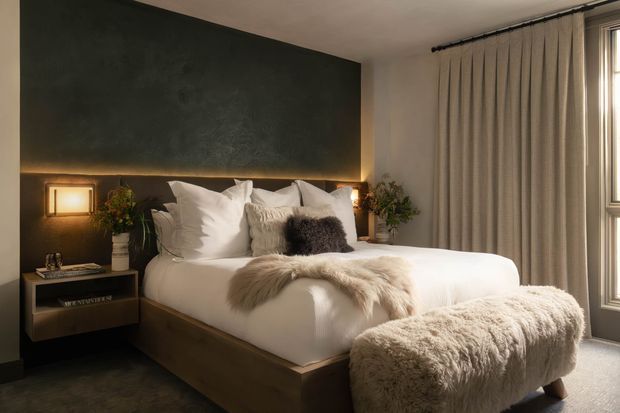 A warm, moody contemporary bedroom featuring a low timber platform bed with layered white bedding, textured throws and pillows, a dark charcoal feature wall with concealed warm lighting, floating bedside joinery, framed wall lights, full-height beige curtains, and a plush upholstered bench at the foot of the bed.