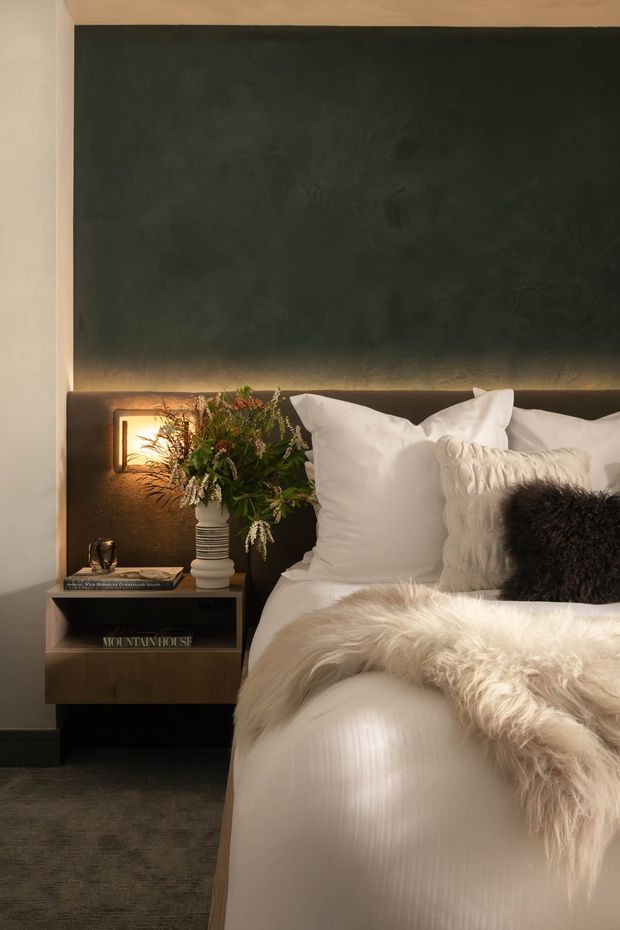 A moody, refined bedroom vignette featuring a bed with layered white bedding, textured cream and dark cushions, and a fluffy light throw. A dark charcoal feature wall with warm integrated bedside lighting sits behind the bed, alongside a floating dark timber bedside table with books, a vase, and greenery. The room has soft, warm ambient illumination and dark carpet flooring.