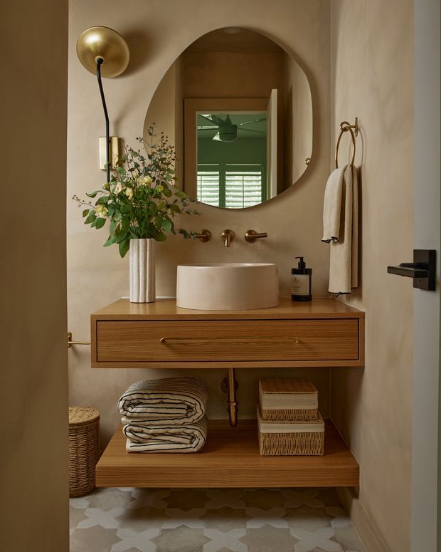 Warm, compact powder room featuring a floating timber vanity with open lower shelving, a round vessel basin, wall-mounted brass tapware, an organically shaped mirror, brass wall sconce and towel rail, and patterned geometric floor tiles. Neutral plaster walls and natural textiles create a refined, cozy atmosphere.