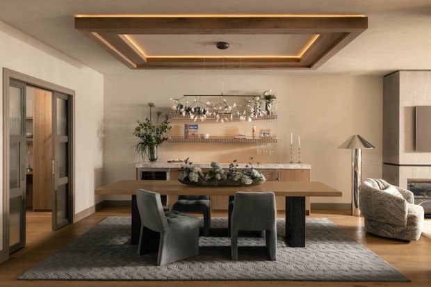Refined dining room with a substantial timber dining table, upholstered dark dining chairs, a large patterned rug, and a long built-in sideboard with open display shelving. Warm integrated lighting highlights the recessed timber ceiling feature, while glazed doors open toward an adjacent room and a sculptural floor lamp and lounge chair sit to the right.