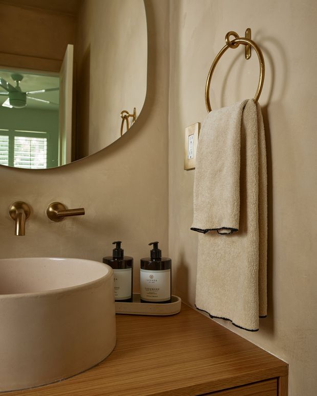Close-up view of a warm, refined bathroom vanity with a round vessel basin on a timber countertop, wall-mounted brushed brass tapware, a large curved-edge mirror, brass towel ring, neutral plastered walls, and neatly arranged soap dispensers. A bedroom or living space is faintly visible in the mirror reflection.