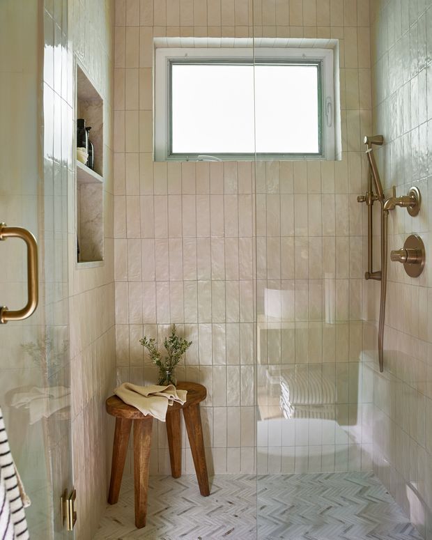 A compact walk-in shower bathroom with warm beige vertically tiled walls, a small horizontal window, recessed shower niche, brushed brass shower fixtures, clear glass shower screen, and a herringbone-patterned light grey floor. A rustic wooden stool with folded towels and greenery adds warmth.