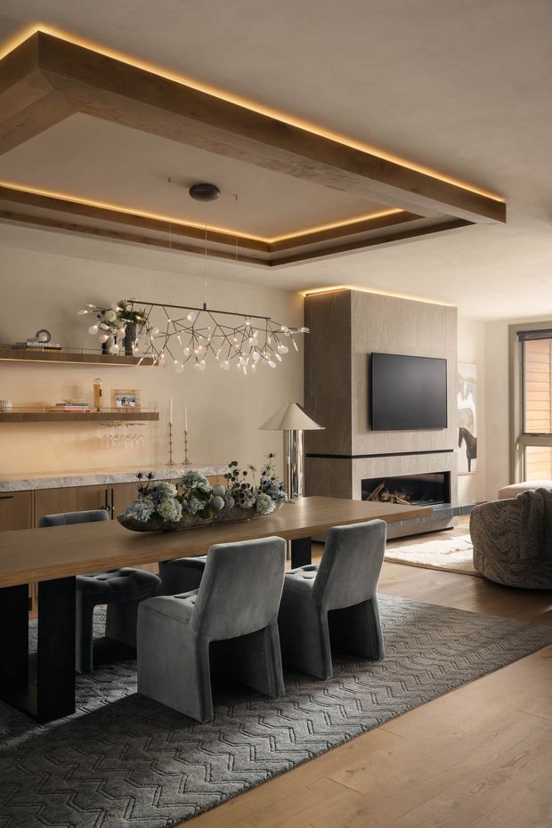 A warm, refined open-plan dining and living area with a long dark timber dining table, upholstered grey dining chairs, built-in shelving, a sculptural pendant light, and a fireplace/media wall. The room features a layered recessed timber ceiling with concealed amber lighting, light wood flooring, a large patterned grey rug, and neutral textured finishes.