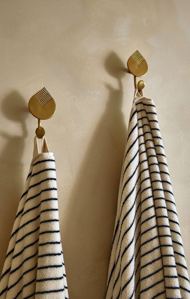 Close-up of two cream-and-black striped fabric towels hanging from decorative brass-gold wall hooks on a warm beige textured wall. The image focuses on wall-mounted bathroom or utility accessories and tactile materials, with strong warm lighting and shadows.