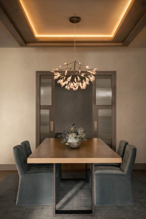 A refined dining room with a rectangular timber dining table, upholstered charcoal dining chairs, and a sculptural branching pendant light. The room features warm concealed cove lighting in a recessed ceiling, neutral plaster walls, dark framed glazed double doors, and dark carpeting. A floral arrangement sits centrally on the table, creating a warm, sophisticated atmosphere.