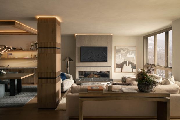 Warm, refined open-plan living space with a contemporary lounge centred on a stone-textured fireplace wall and wall-mounted television. A large neutral sectional sofa faces a substantial timber coffee table, while a sculptural timber-clad column separates the living area from a dining and kitchen zone with open shelving and integrated lighting. Expansive corner glazing brings in natural light and views of trees.