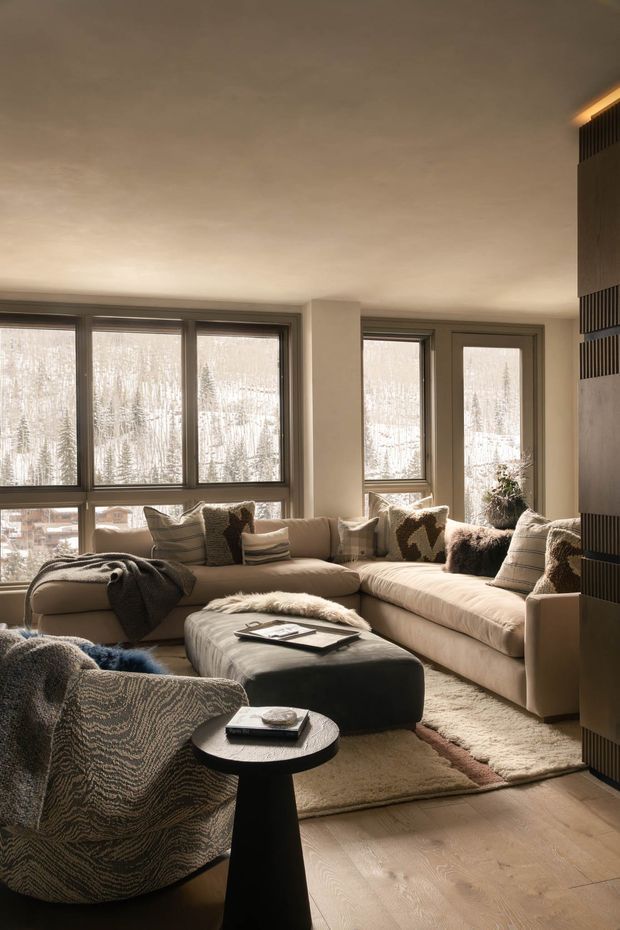 Warm, softly lit contemporary living room with large windows overlooking a snowy landscape. The room features a generous cream sectional sofa, textured neutral cushions, a dark upholstered ottoman, a sculptural black side table, a patterned armchair, a pale textured rug, light timber flooring, and a dark vertical slatted feature wall or divider. The styling is cozy and refined with layered textiles and natural neutral tones.