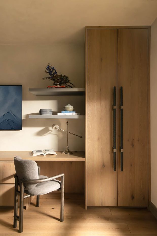 A warm, compact home office with a built-in timber desk, open shelving, tall timber wardrobe or storage cabinetry, upholstered armchair, desk lamp, books, and understated decorative objects. Strong natural sunlight creates pronounced shadows across the neutral walls and timber floor.