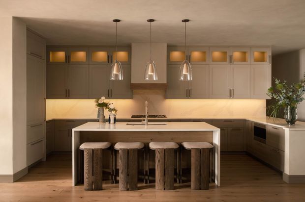 Warm, refined kitchen featuring a large central island with four rustic timber stools, extensive pale panelled cabinetry, integrated warm under-cabinet lighting, a stone-look benchtop, and three pendant lights. Dark timber flooring and floral arrangements add contrast to the neutral scheme.