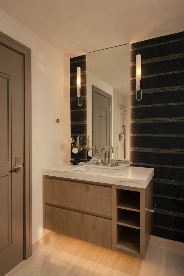 A refined bathroom vanity area with a warm timber floating cabinet, white marble benchtop, integrated basin, polished metal tapware, and open shelving. A large mirror is framed by dramatic dark horizontally textured wall panels with vertical illuminated sconces. The room also includes a paneled grey door, light timber flooring, and a glimpse of a glass shower enclosure in the mirror.