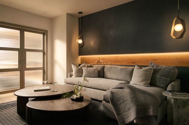 A warm, moody living room with a large neutral sectional sofa, layered cushions and throw, dark textured feature wall, and a long illuminated timber wall shelf or ledge behind the sofa. Two low dark coffee tables sit on a patterned rug, while pendant lights and glazed doors provide soft ambient light.