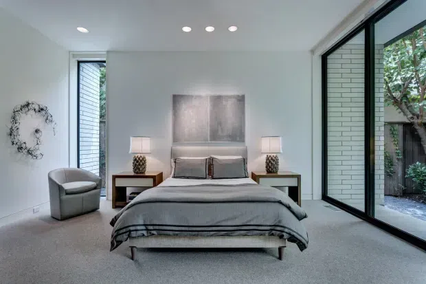A bright contemporary bedroom with a low upholstered bed centered against a white wall, flanked by matching dark bedside tables and lamps. Floor-to-ceiling black-framed glazing opens views to a planted courtyard, while a grey armchair, pale carpet, abstract wall art, and soft grey bedding create a calm, refined interior. Recessed ceiling lights and clean-lined detailing reinforce the modern architectural character.