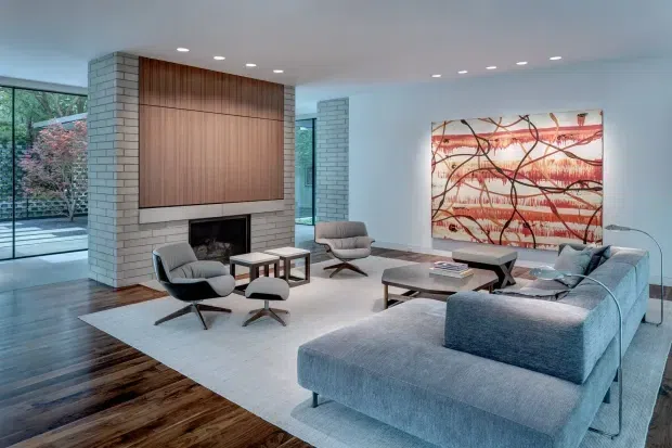 A spacious, light-filled contemporary living room with a large grey sectional sofa, sculptural lounge chairs, low coffee tables, and a pale area rug. A white-painted brick fireplace wall is paired with a full-height warm timber feature panel and dark firebox. Floor-to-ceiling glazing opens to a landscaped exterior, while a large abstract artwork provides a red and black accent.