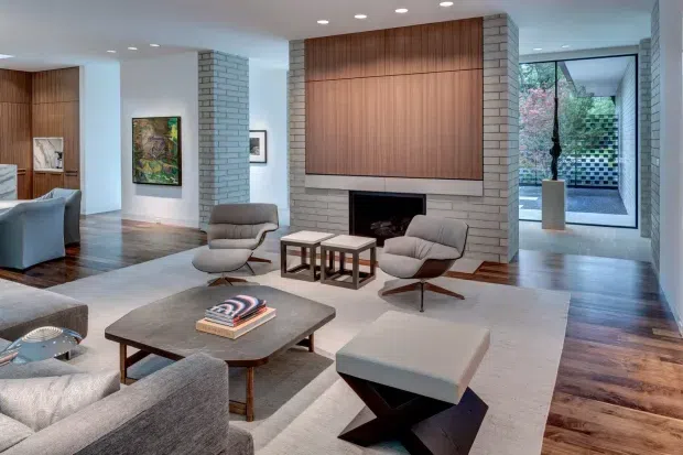 A refined, spacious mid-century-inspired living room with a large light rug, sculptural lounge chairs, upholstered seating, a low dark coffee table, and a substantial fireplace wall. The fireplace is framed by pale grey brick columns and a vertical timber-panelled feature wall, with recessed ceiling lighting and views toward a glazed courtyard. Dark timber flooring continues through the open-plan space.