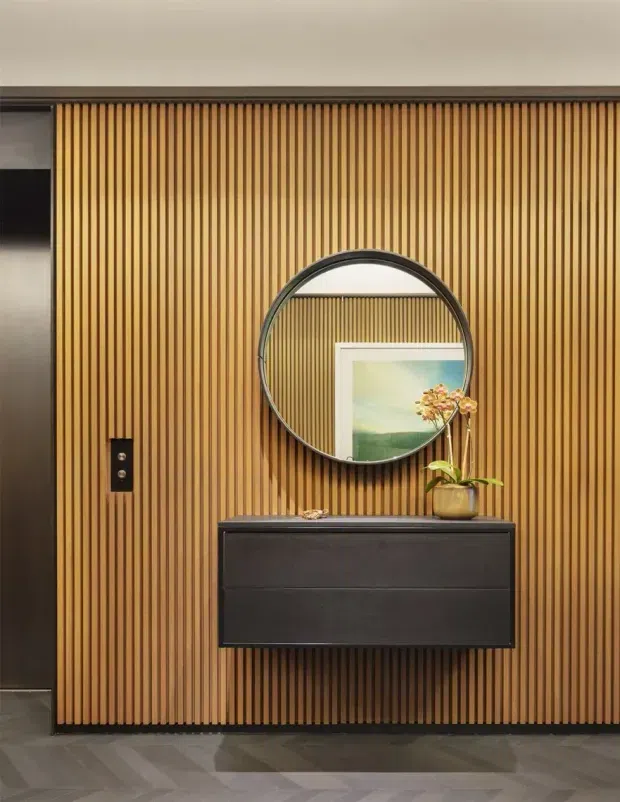 A refined interior entryway featuring a full-height vertical timber slat feature wall in warm honey tones. A round black-framed mirror hangs above a sleek dark floating console, with a small potted orchid and framed artwork reflected in the mirror. Dark tiled flooring and a concealed dark doorway or opening frame the composition. The look is clean, warm, and contemporary with strong linear texture and contrast.