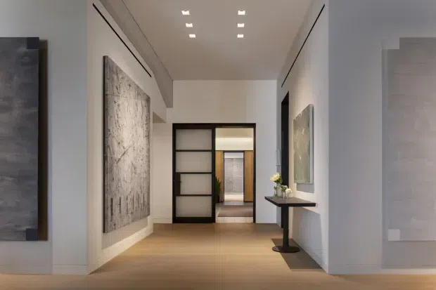 A refined modern hallway or entry corridor with warm timber flooring, white walls, recessed linear ceiling lighting, and black-framed glazed sliding doors opening toward another room. Large monochromatic wall art and a slim black console table accent the space. The composition emphasises clean lines, built-in architectural detailing, and a calm neutral palette.
