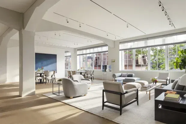 Bright, spacious contemporary living room in a loft-like interior, with white structural columns and beams, high ceilings, expansive industrial-style multi-pane windows, and track lighting. The room contains multiple upholstered lounge chairs and sofas, coffee tables, a large pale rug, and a small dining area near a dark feature wall. Warm timber flooring and abundant daylight create an airy, refined atmosphere.