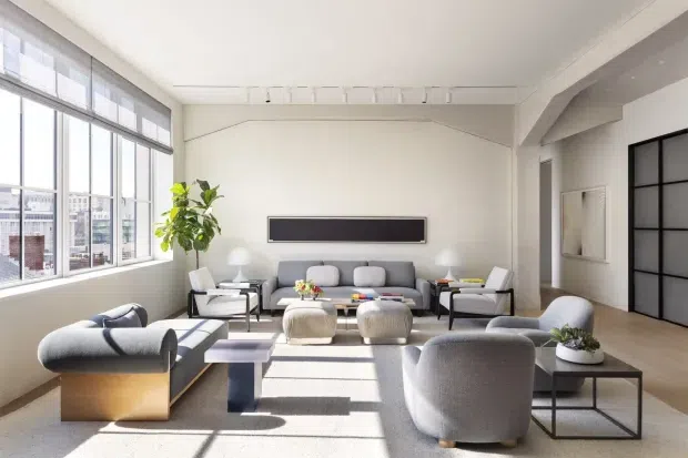Bright, spacious contemporary living room with large industrial-style windows, pale walls and flooring, a grey sofa and rounded lounge chairs arranged around a low coffee table, and a large black-framed glazed sliding door. Strong daylight creates a calm, refined atmosphere.