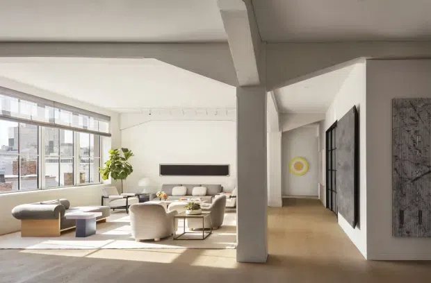 Bright, spacious contemporary living room with high ceilings, large industrial-style multi-pane windows, pale timber flooring, and a neutral seating arrangement. A substantial structural column and beam divide the living area from a hallway, while abstract wall art, a potted indoor tree, and a wall-mounted linear fireplace add visual interest. Urban buildings are visible through the glazing.