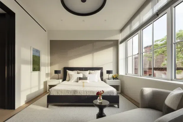 A bright, contemporary bedroom with a low-profile dark bed, layered white bedding, matching bedside tables and lamps, and a grey upholstered lounge chair in the foreground. Full-height windows line one side of the room, bringing in abundant natural light. The space has pale walls, a light carpet, dark timber-toned accents, a large round ceiling light, and restrained modern styling.