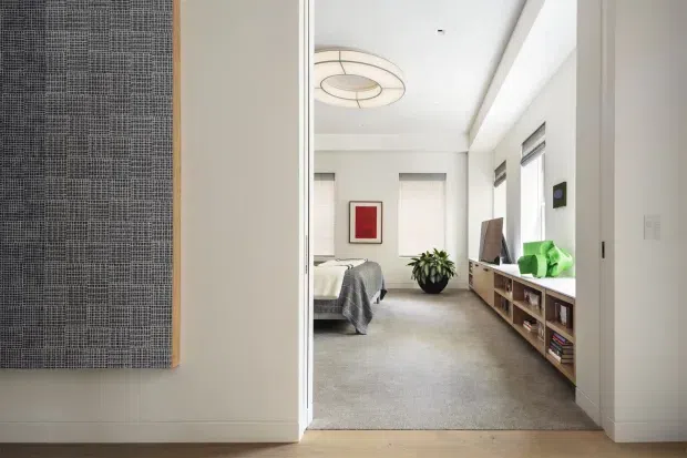 View through a wide doorway into a bright, minimalist bedroom and adjoining living area. The room features pale carpet, white walls and ceiling, a low timber media/storage console beneath windows, a grey upholstered bed or seating area, a potted plant, and a simple circular pendant light. A textured monochrome wall artwork is visible in the foreground.
