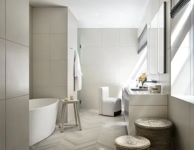 Bright contemporary bathroom with a freestanding white bathtub, wall-mounted double vanity, large mirrors, white tiled walls, and a pale herringbone tile floor. A compact upholstered stool and woven round stools add subtle texture, while high windows with blinds bring in strong natural light.
