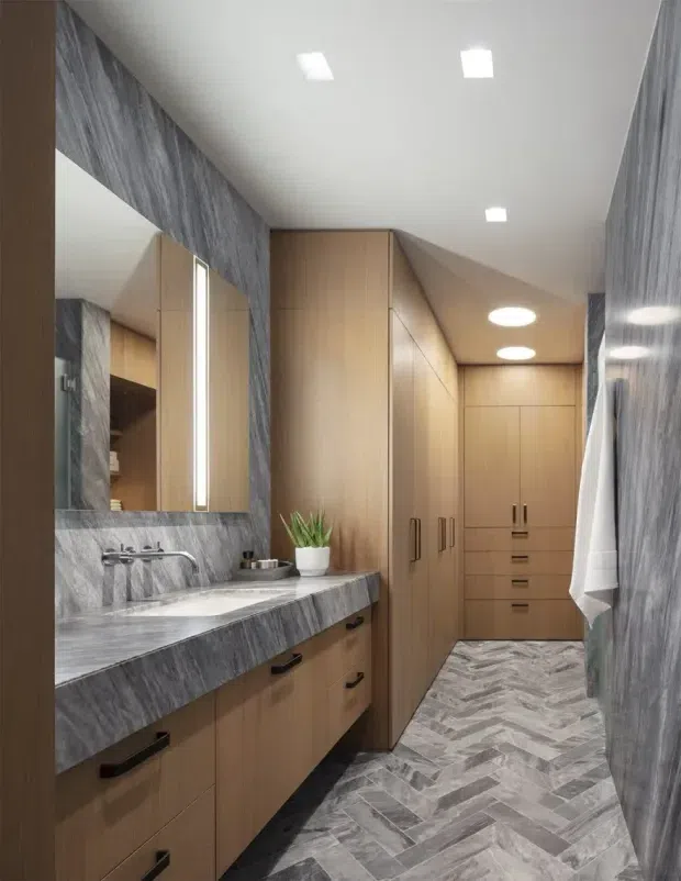 A narrow, modern bathroom with a long grey marble-look vanity and integrated basin, wall-mounted chrome tapware, a large mirrored cabinet, and extensive warm timber cabinetry. The space includes tall built-in storage at the far end, grey herringbone-patterned floor tiles, grey stone-look wall surfaces, recessed ceiling lights, and a white towel hanging on the right.