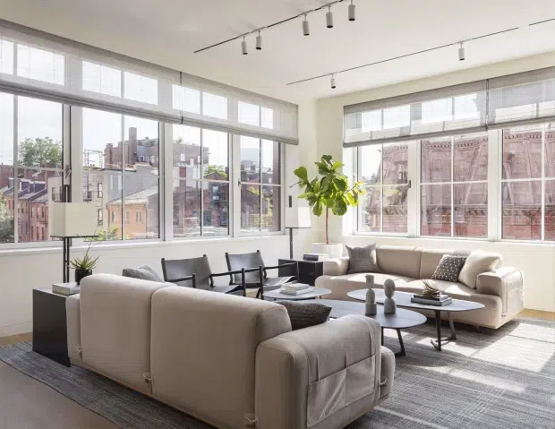 Bright, spacious urban living room with expansive industrial-style windows, roller shades, a large neutral sectional sofa, matching armchairs, layered coffee tables, and a contemporary track-lighting system. The room overlooks dense city buildings and is softened by indoor plants, textured cushions, and a large area rug.