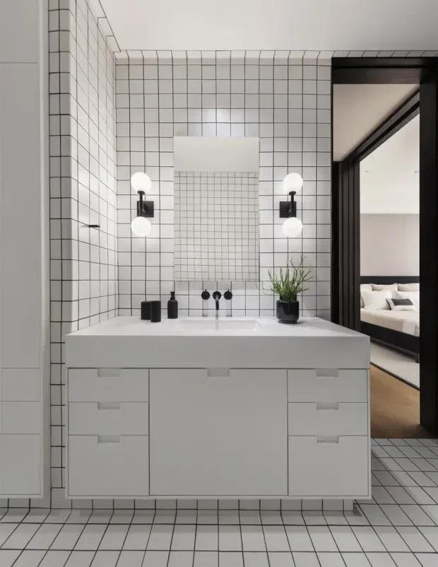 A contemporary bathroom centred on a floating white vanity with integrated basin, black tapware, and a rectangular mirror. Small white square tiles with dark grout cover the walls and floor, creating a strong grid pattern. Globe wall sconces flank the mirror, while a potted plant and black accessories add contrast. A dark-framed opening reveals part of an adjacent bedroom.