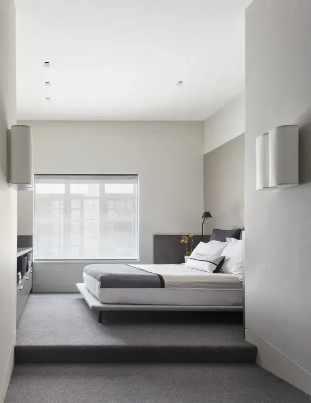 A minimalist bedroom viewed from a slightly lower, carpeted level. The room has a low platform bed with white bedding and charcoal-grey accents, a dark upholstered headboard, integrated bedside lighting, pale grey walls, recessed ceiling spotlights, built-in cabinetry, and a large window with a translucent blind. The bedroom is separated from the foreground by a broad raised carpeted platform step.