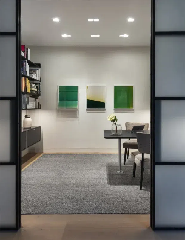 A refined, compact home office or study viewed through a dark-framed glazed doorway. The room has pale walls, a white recessed ceiling with downlights, built-in dark shelving and cabinetry along the left wall, three green-toned artworks, a small black table with chairs, and a large grey area rug over timber flooring. The scheme is restrained and contemporary with strong black, white, grey, and green contrasts.