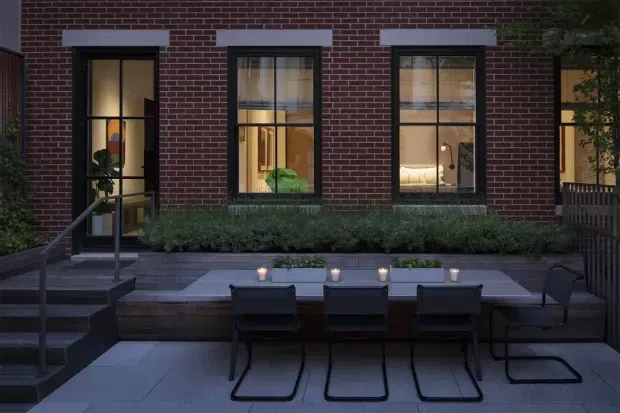 A dusk view of a refined urban courtyard terrace adjoining a dark red brick building. Black-framed multi-pane windows and a glazed door reveal warmly lit interiors, while a long built-in dining table with black chairs, planters, low planting, steps, and dark metal railings define the outdoor space.