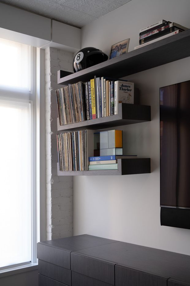 A refined contemporary living-room media wall with three dark timber floating shelves displaying books, vinyl records, framed photographs and decorative objects. A large wall-mounted television is partially visible at right above a matching dark timber storage cabinet. The room has a pale neutral wall, exposed painted brick at the window reveal, a patterned pressed-tin ceiling, and tall sheer-covered glazing.