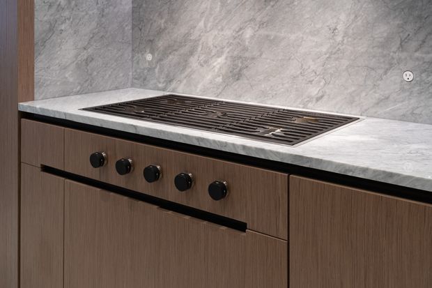 Close-up view of a contemporary kitchen cooking zone with a large integrated gas cooktop set into a pale veined stone benchtop. The splashback is clad in matching grey marble-look stone, while the lower cabinetry has dark warm wood-grain fronts with black handles and controls.