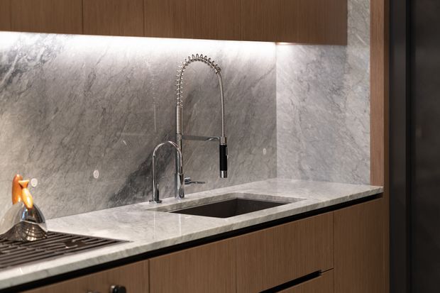 Close-up of a contemporary kitchen workstation featuring a grey veined stone benchtop and matching full-height splashback, an undermount sink with a spring-coil pull-down faucet, and warm timber-grain cabinetry. Integrated under-cabinet lighting highlights the stone surfaces.