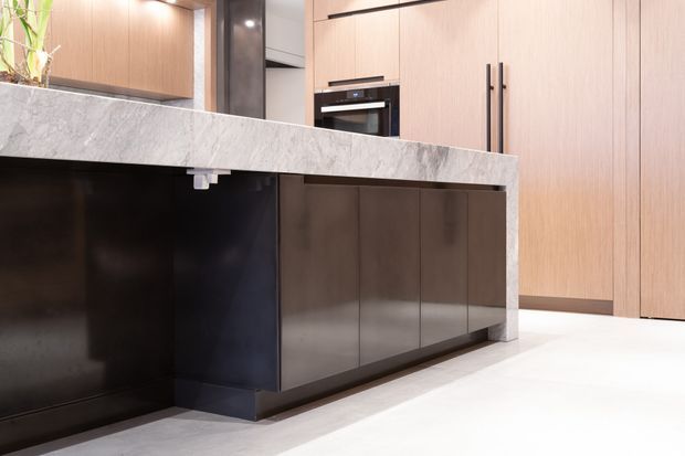 Contemporary kitchen with a large island clad in a white-grey veined stone benchtop and waterfall end panel. The island features dark cabinetry and a black open void beneath the overhang, while the surrounding full-height cabinetry is finished in warm light timber with long black vertical handles. Integrated appliances and pale flooring complete the refined, minimalist interior.