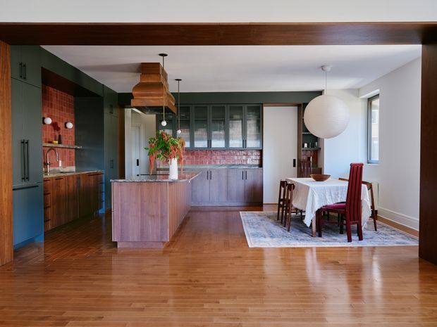 Open-plan kitchen and dining area with warm timber flooring and ceiling beam, dark green cabinetry, timber island and cabinetry, exposed red brick splashback, copper range hood, and a dining table beneath an oversized spherical pendant light. The room combines mid-century-inspired joinery with contemporary detailing and abundant natural light.