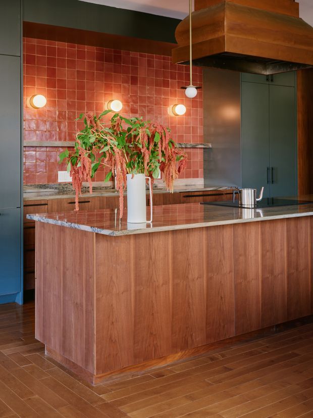 A richly coloured kitchen centred on a large timber island with a grey stone benchtop. The island includes integrated cabinetry, while the rear wall features glossy terracotta-red square tile splashback, timber cabinetry, dark green full-height joinery, and a substantial copper-toned rangehood. Globe wall and pendant lights provide warm illumination, and a potted plant adds organic texture.