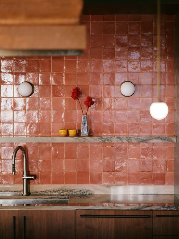 Close-up view of a richly styled kitchen featuring glossy terracotta-pink square wall tiles, a dark timber rangehood and cabinetry, a stone benchtop, and a tall brushed metal gooseneck tap. Two round wall lights and a small decorative vase add warm, refined character.