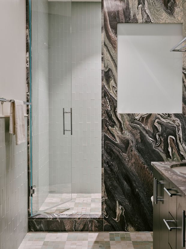 A sophisticated contemporary bathroom featuring a glass-enclosed walk-in shower with pale vertically arranged wall tiles, a small-format mosaic shower floor, and a striking dark veined stone feature wall surrounding a large frameless mirror. A matching stone vanity benchtop and dark cabinetry are visible at right, with chrome hardware and towel rails.
