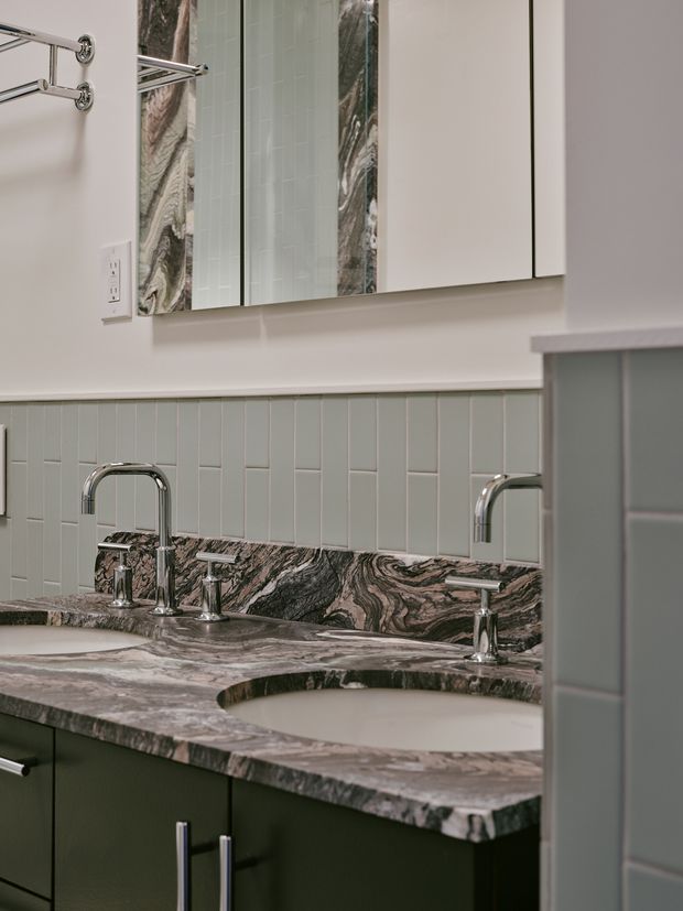 Close-up of a refined double-vanity bathroom with two undermount basins, polished chrome gooseneck taps, a heavily veined natural stone benchtop and matching backsplash, pale blue-green vertical wall tiles, dark green cabinetry, and mirrored wall cabinets.