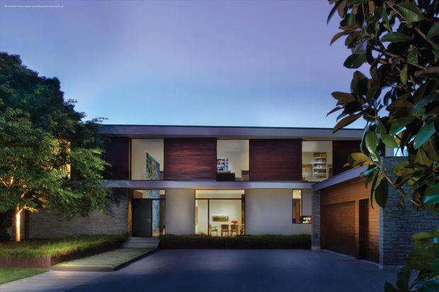 Twilight exterior view of a sophisticated two-storey contemporary residence, framed by mature landscaping. The home has a strong horizontal composition with a flat roof, dark masonry walls, warm timber-clad garage and upper-level panels, recessed glazing, and a sheltered entry approached by a paved driveway and steps. Warm interior lighting contrasts with the cool blue dusk sky.