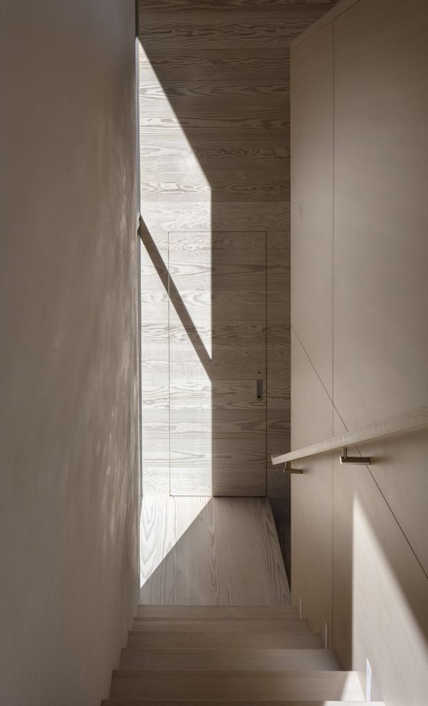 A narrow, minimalist stairway is enclosed by pale timber and smooth light-coloured walls. The stair treads descend toward a small landing with a flush timber door, while built-in cabinetry and a simple timber handrail line the right wall. Strong sunlight casts dramatic geometric shadows across the wood-grain surfaces, emphasizing the vertical enclosure and refined detailing.