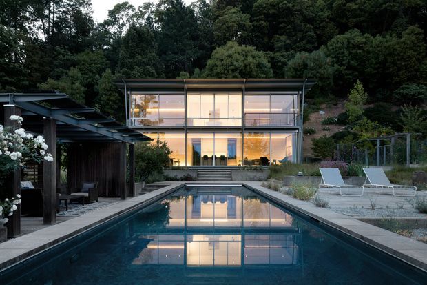 A contemporary multi-level glass-fronted house set into a lush wooded hillside, viewed from a long rectangular swimming pool. The residence features extensive floor-to-ceiling glazing, dark structural framing, projecting rooflines and warm interior illumination. A covered outdoor entertaining pavilion sits to the left, while pale lounge chairs and landscaped planting surround the pool deck.