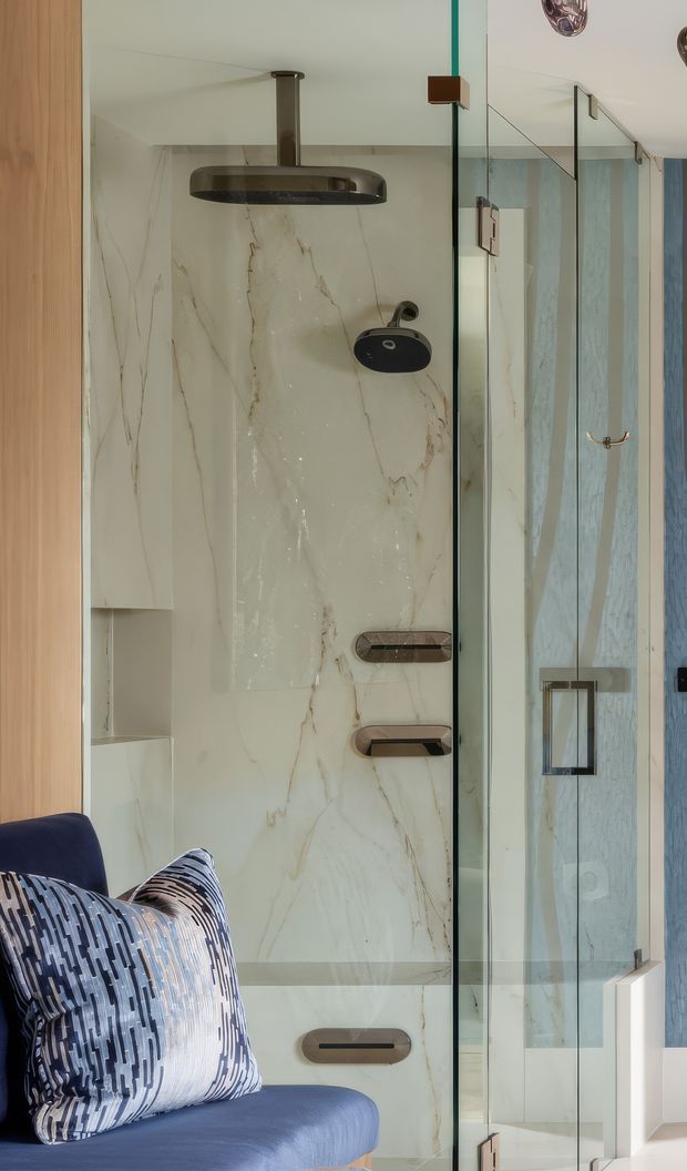 A refined bathroom shower area viewed beside a blue upholstered chair. The shower is enclosed by clear frameless glass with dark metal hardware, a large cream marble-look wall and floor surface with subtle veining, and matching dark bronze or black shower fixtures. Built-in white shelving is visible at the left, while a mirrored or glazed surface reflects the bathroom and adjacent blue-toned wall details. The overall composition is luxurious and contemporary with a restrained neutral and blue palette.